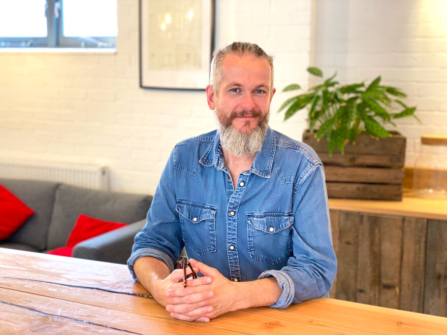 Nik smiling and sat at a table with his glasses in his hands.