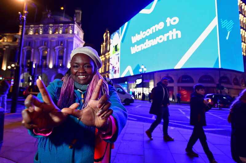 A woman poses with her hands in a 'W' shape in front of the Piccadilly Screen