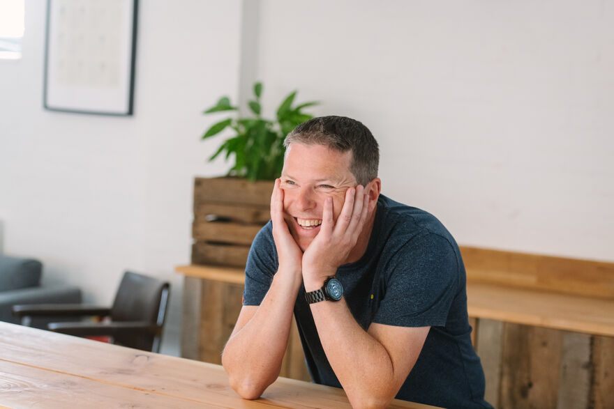 Sam sat at a table and smiling with his head resting in his hands.
