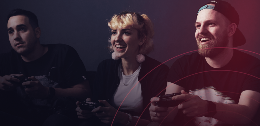 Three people holding console contollers and smiling while playing a game with a pink, dark blue overlay and pink circles coming from the bottom right. The bottom left has the studio gobo logo