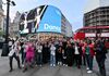 A large group of people make a 'W' symbol with their hands in front of the Piccadilly screen.
