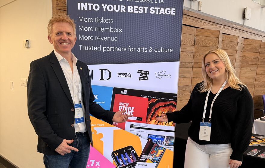 Paul and Danie at a conference stood in front of a stand that reads 