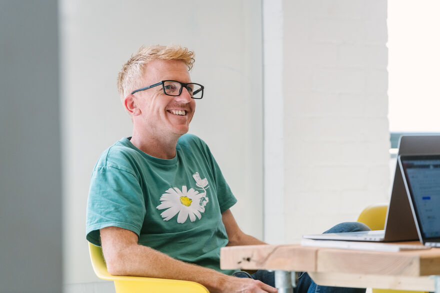Paul sat in a yellow chair at a table and smiling while looking at his laptop.