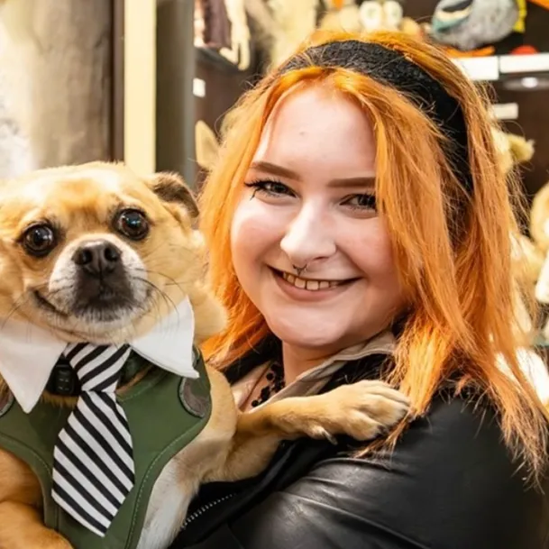 Ajna, a white woman with ginger hair smiling while holding a small ginger dog which is smiling and wearing a black and white stripy tie and a white collar. 