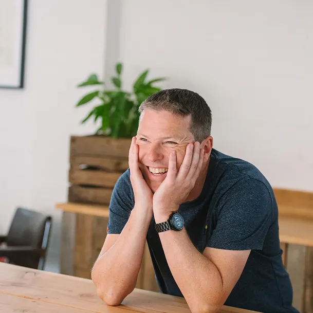 A headshot of Sam, a white man with short, dark hair, he is smiling and resting his head in his hands.