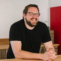 A headshot of Vince, a white man with dark hair, glasses and a beard, smiling with his hands rested on the table.
