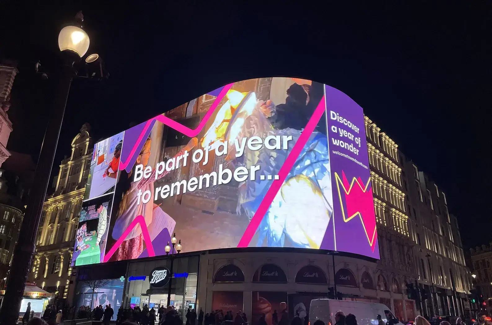 Looking up at the Piccadilly screen, it is showing a W mark over an image with text reading Be part of a year to remember….