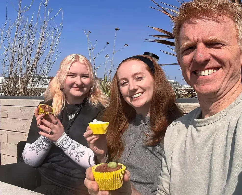 Paul, Carrigan and Ajna take a selfie in a rooftop garden in Brighton, they are each holding cupcakes. 