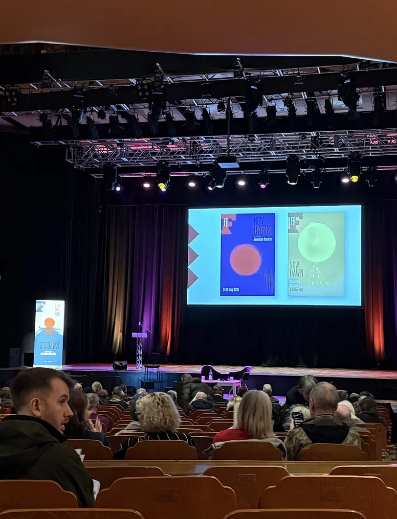 Looking up at a stage from the perspective of the audience, behind the stage is a large screen displaying a picture of Brighton Festival posters.
