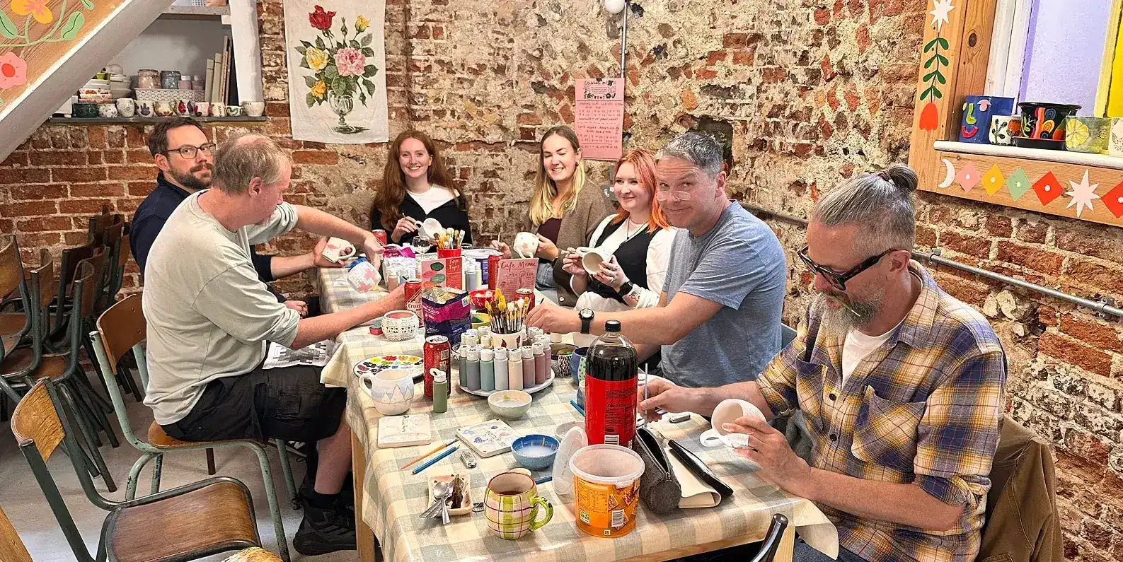 The Grandad team sit at a long table in a room with flint walls, painting mugs. 