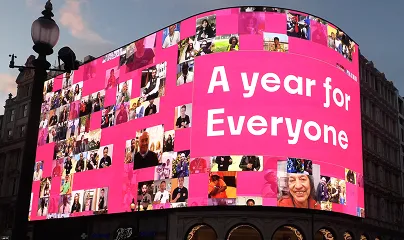 Looking up at the Piccadilly Screen which has a pink background, a collage of pictures of happy people and text reading “A year for everyone”